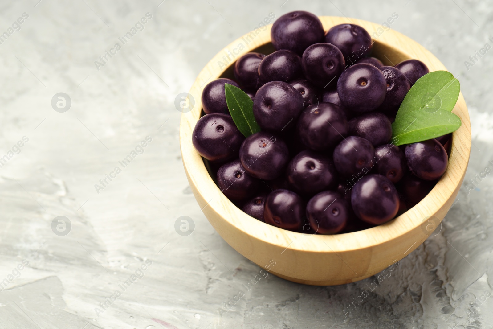 Photo of Ripe acai berries and leaves in bowl on grey textured table, closeup. Space for text