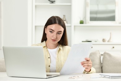 Budget planning. Young woman with papers using laptop at white table indoors