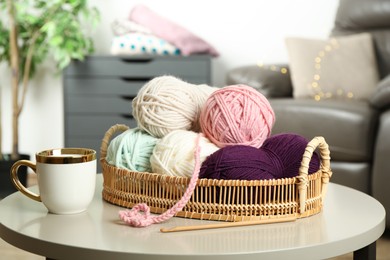 Photo of Wicker basket with colorful yarns, crochet pattern, hook and cup on coffee table indoors