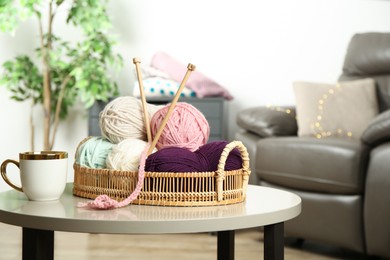 Photo of Wicker basket with colorful yarns, crochet pattern, knitting needles and cup on coffee table indoors