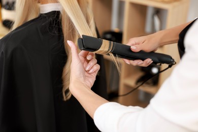 Photo of Hairdresser curling woman's hair with flat iron in salon, closeup