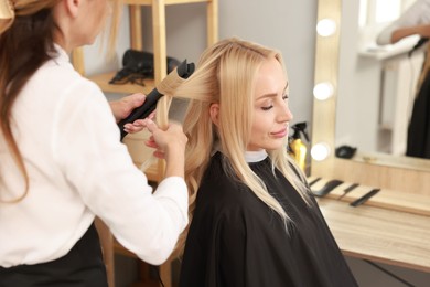 Photo of Hairdresser curling woman's hair with flat iron in salon, closeup