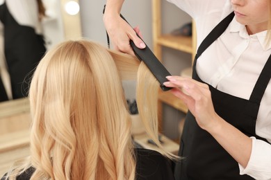 Photo of Hairdresser curling woman's hair with flat iron in salon, closeup