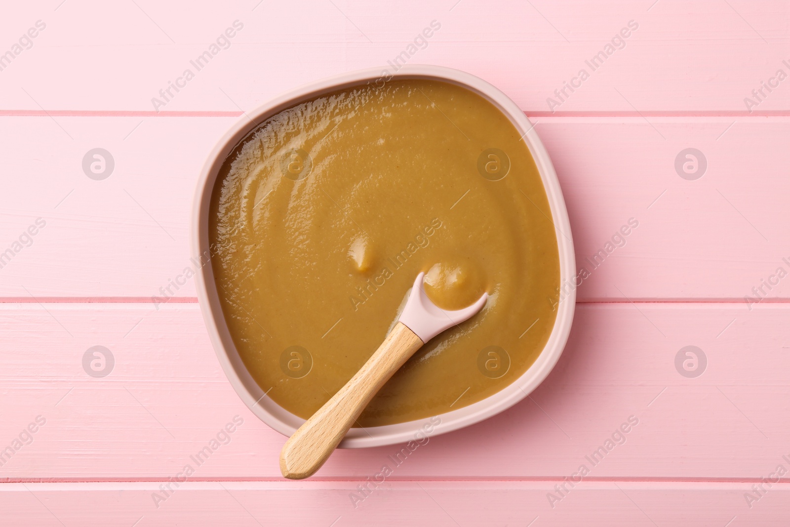 Photo of Delicious baby food and spoon in bowl on pink wooden table, top view