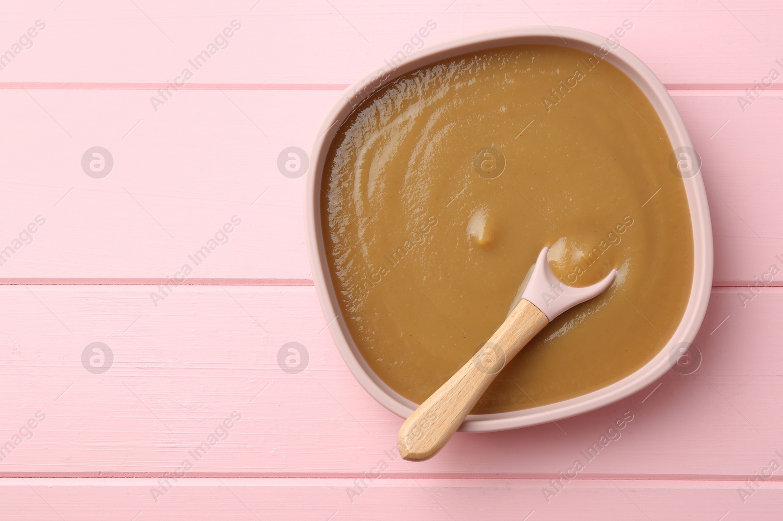 Photo of Delicious baby food and spoon in bowl on pink wooden table, top view. Space for text