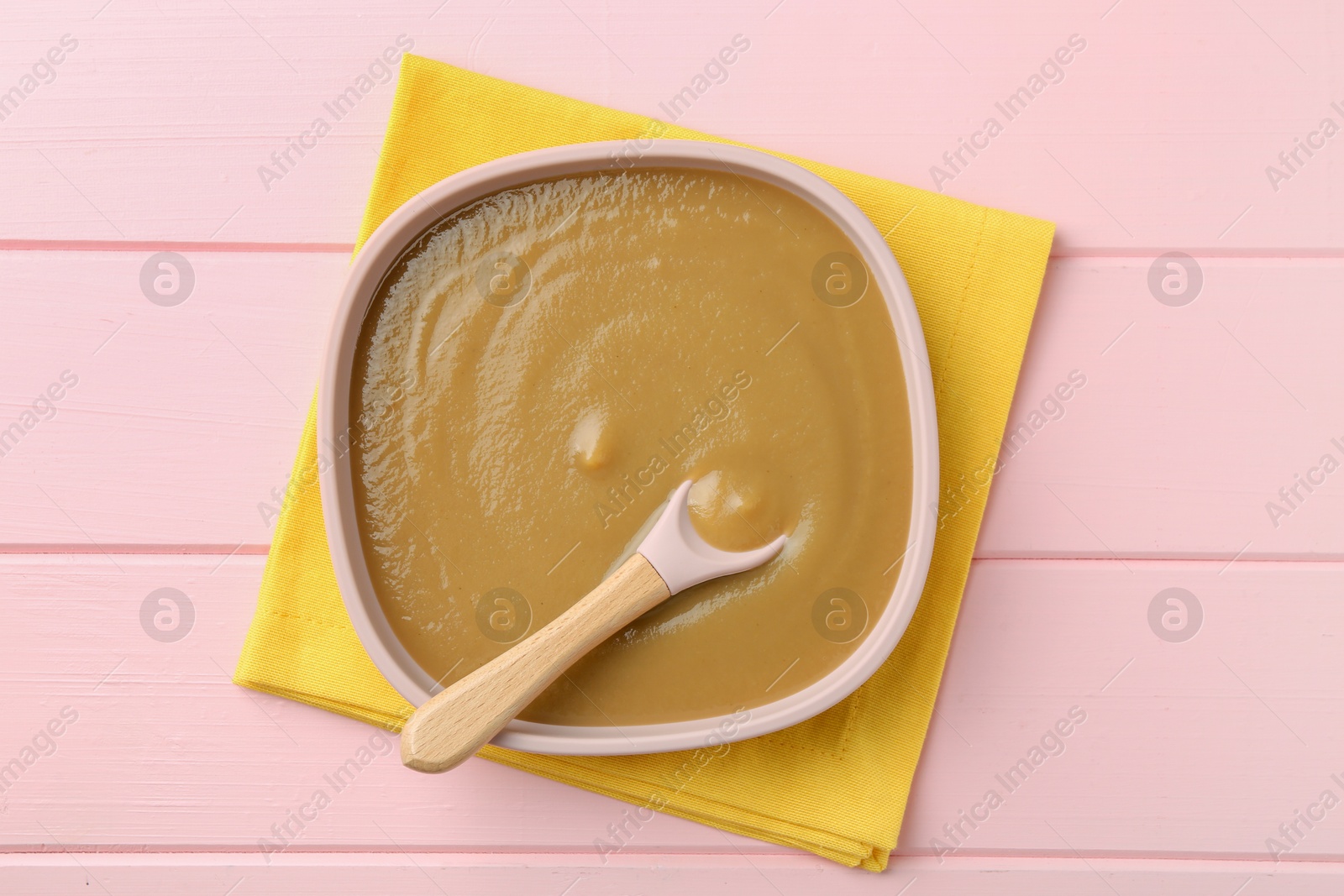 Photo of Delicious baby food and spoon in bowl on pink wooden table, top view