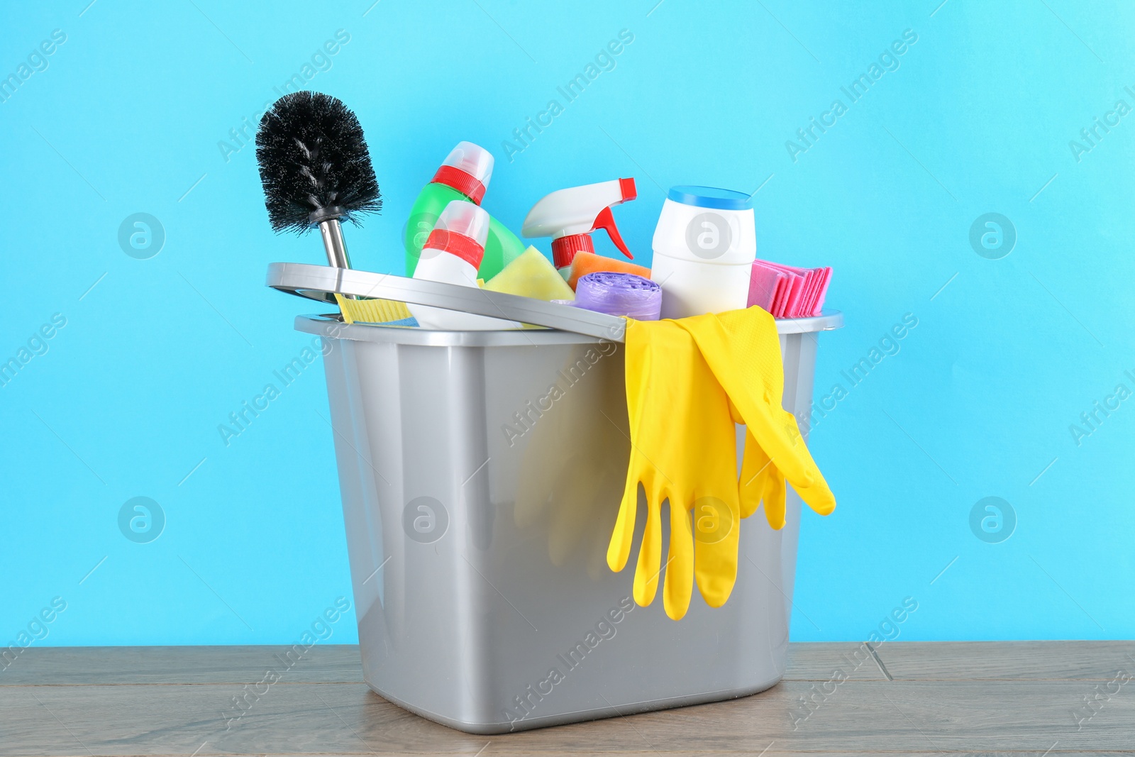 Photo of Bucket with different toilet cleaners, rags, rubber gloves and trash bags on wooden table against light blue background