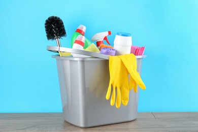 Photo of Bucket with different toilet cleaners, rags, rubber gloves and trash bags on wooden table against light blue background