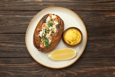 Photo of Tasty baked sweet potato with feta cheese, parsley, sauce and lemon slice on wooden table, top view