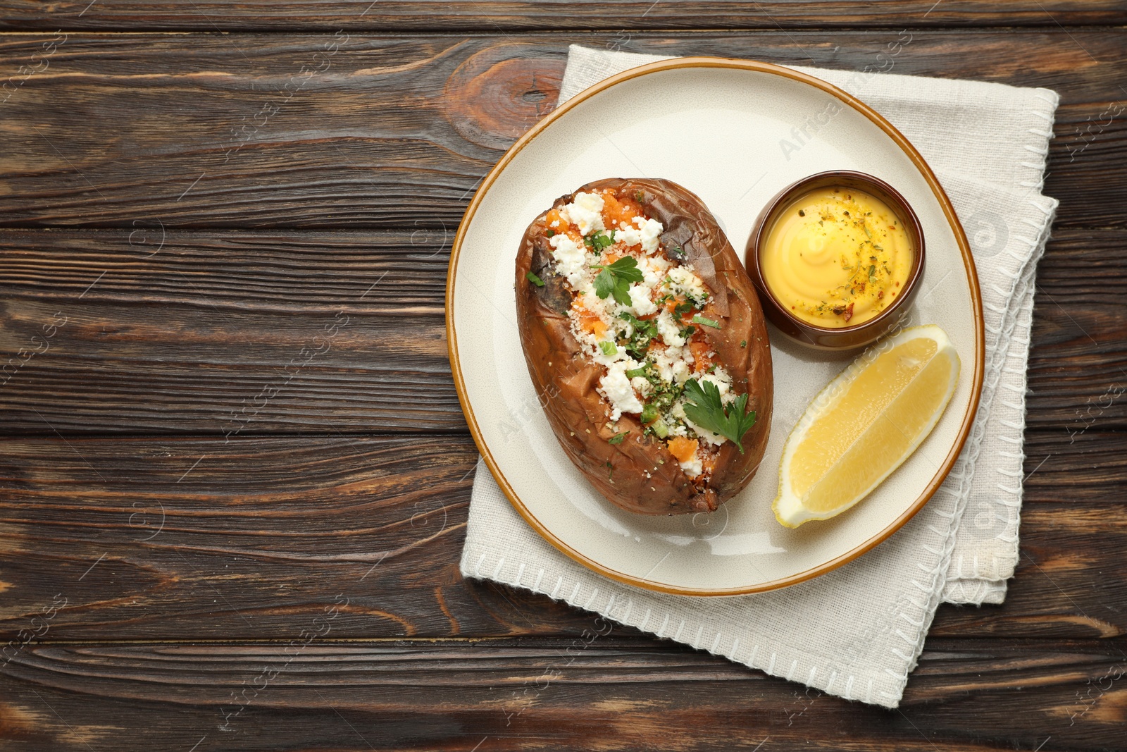 Photo of Tasty baked sweet potato with feta cheese, parsley, sauce and lemon slice on wooden table, top view. Space for text