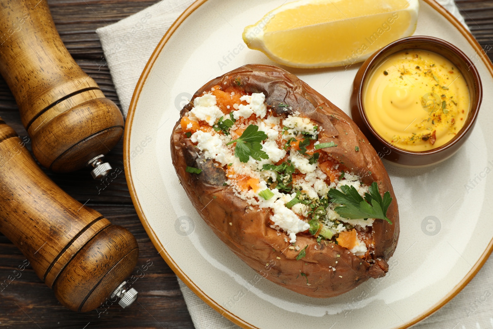 Photo of Tasty baked sweet potato with feta cheese, parsley, sauce, lemon slice and spices on wooden table, flat lay