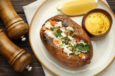 Photo of Tasty baked sweet potato with feta cheese, parsley, sauce, lemon slice and spices on wooden table, flat lay