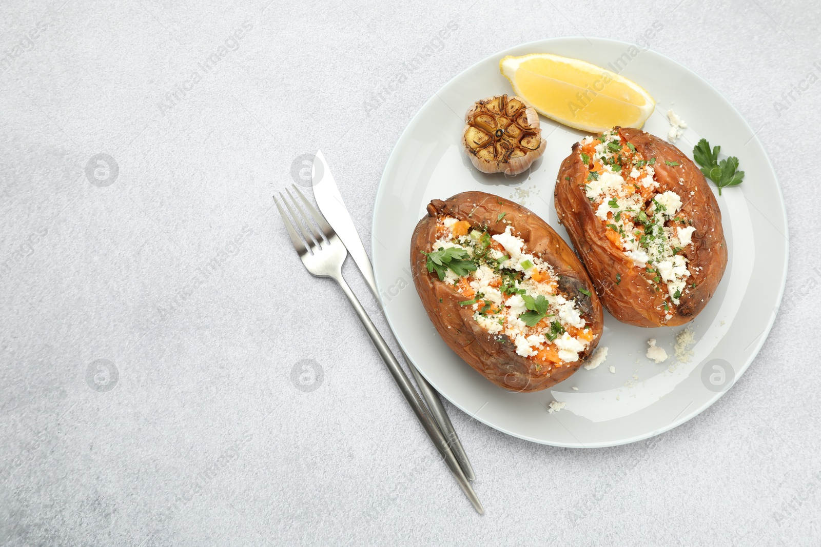 Photo of Tasty baked sweet potatoes with feta cheese, parsley, garlic and lemon slice on light textured table, top view. Space for text