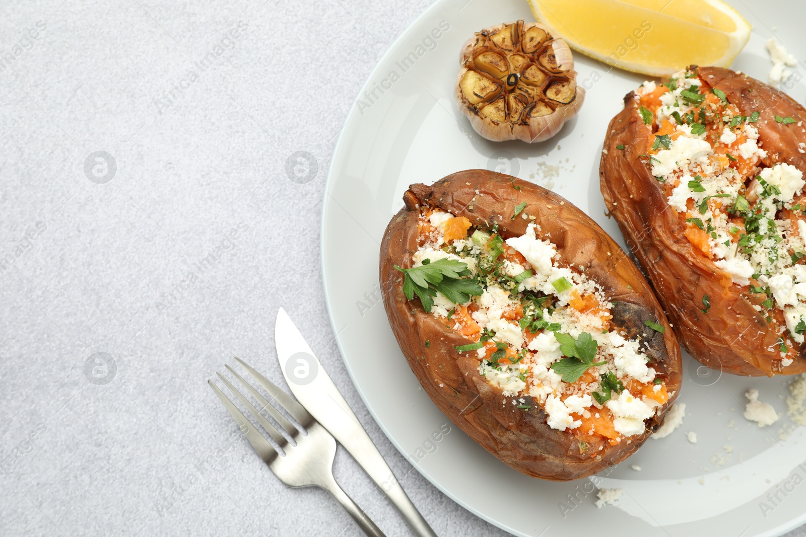 Photo of Tasty baked sweet potatoes with feta cheese, parsley, garlic and lemon slice on light textured table, top view. Space for text