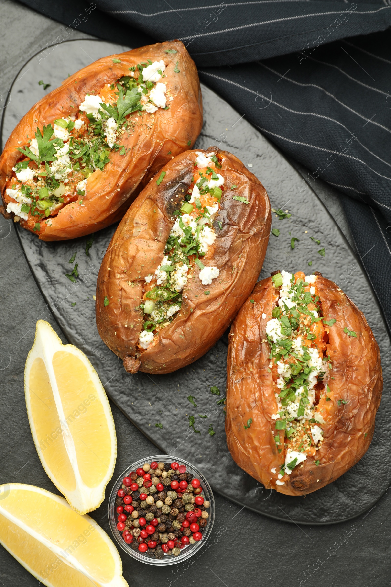 Photo of Tasty baked sweet potato with feta cheese, lemon, peppercorns and herbs on dark textured table, flat lay