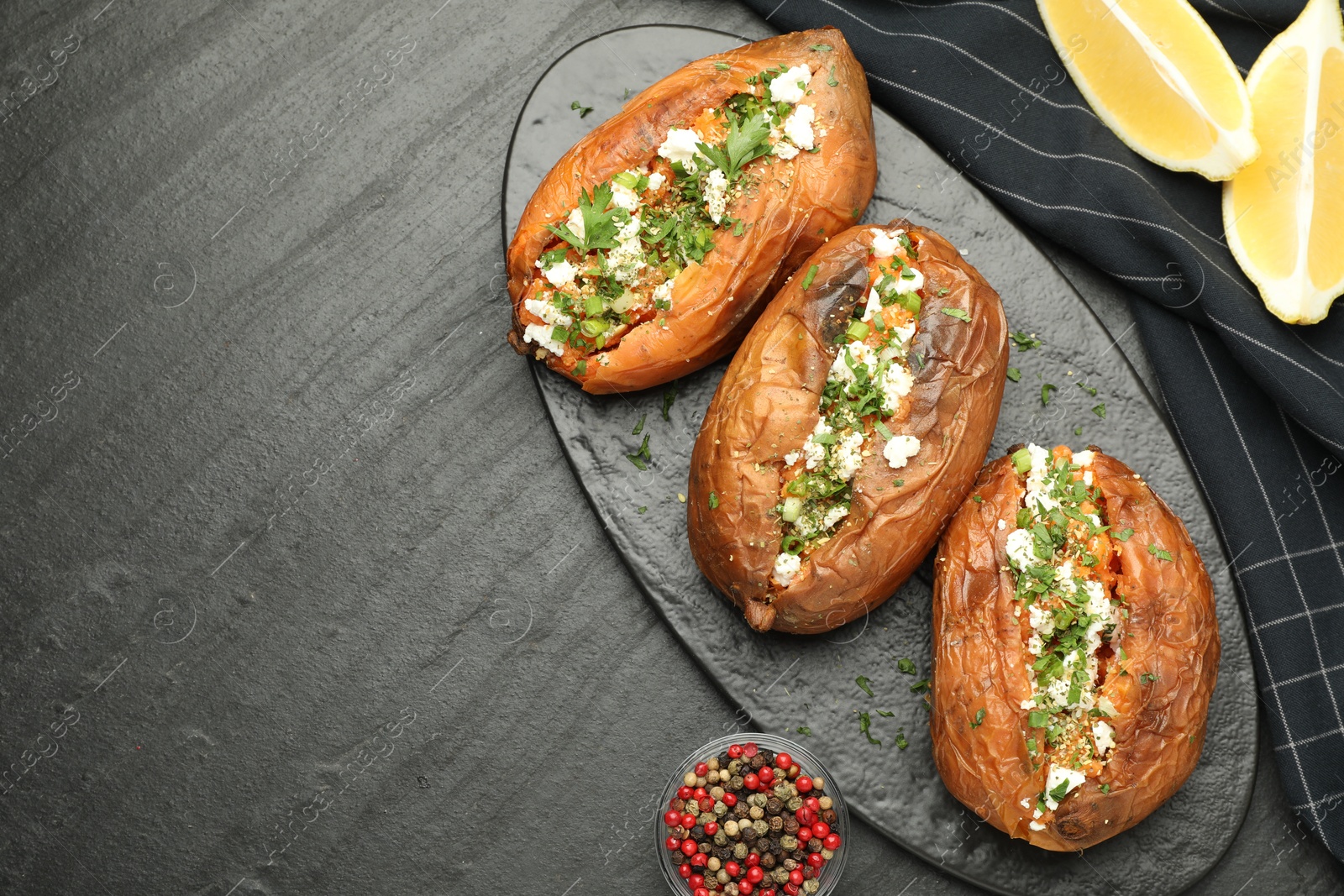 Photo of Tasty baked sweet potato with feta cheese, lemon, peppercorns and herbs on dark textured table, flat lay. Space for text