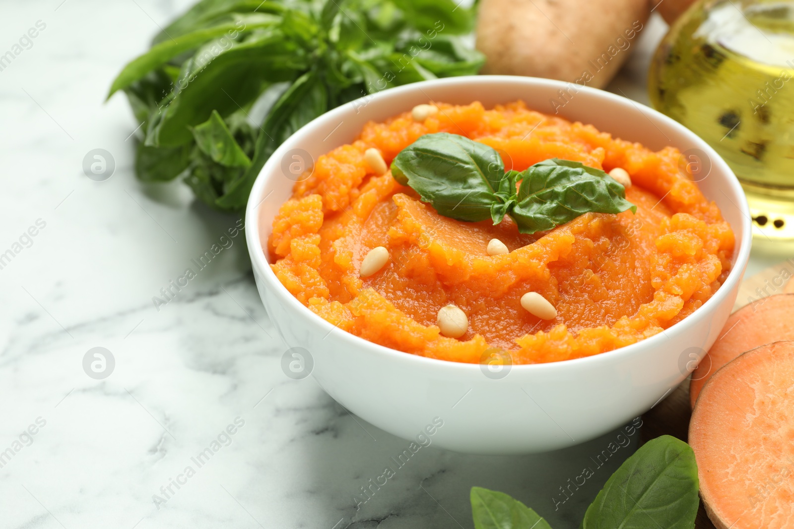 Photo of Tasty mashed sweet potato with basil, seeds in bowl and ingredients on white marble table, closeup