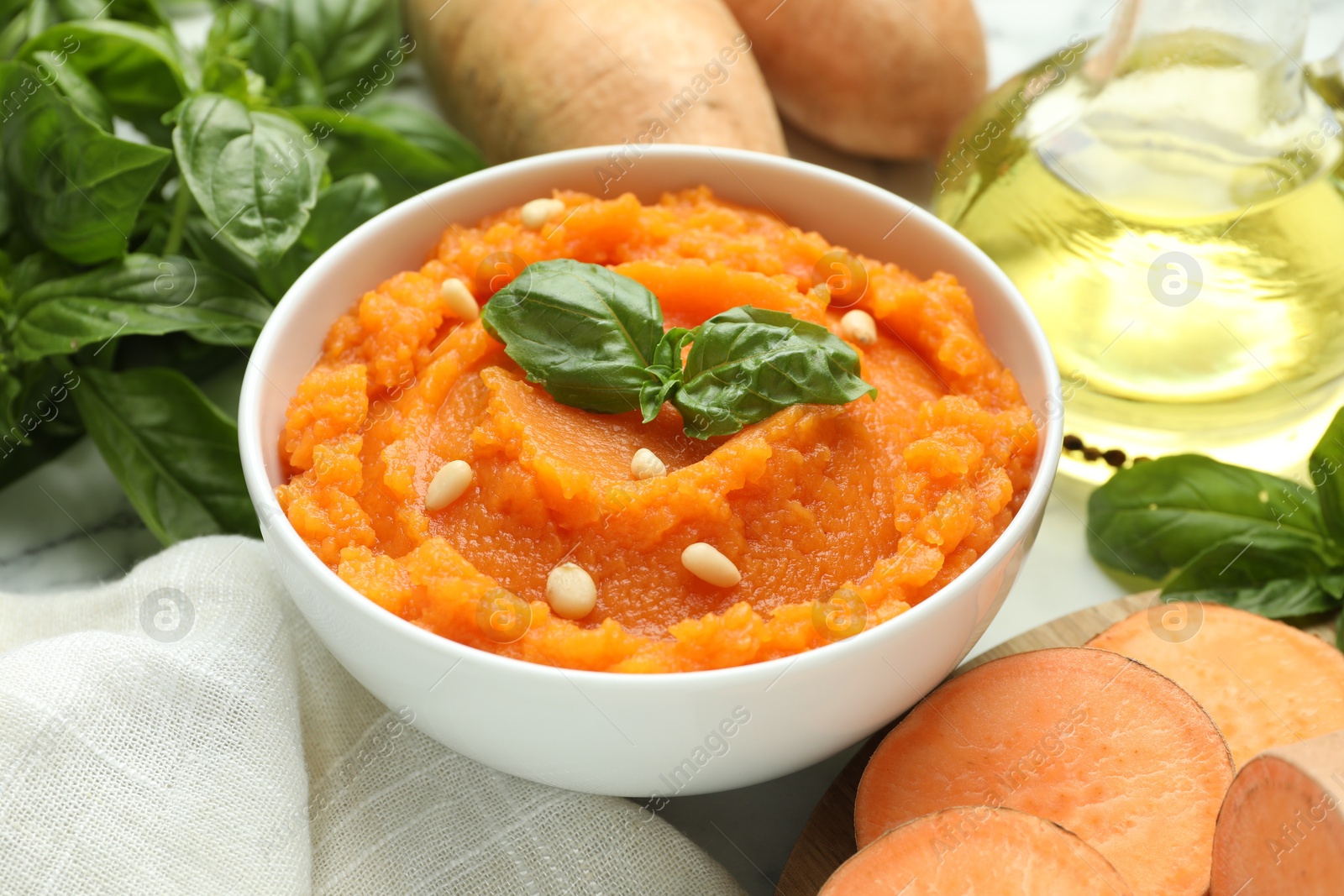 Photo of Tasty mashed sweet potato with basil, seeds in bowl and ingredients on white table, closeup