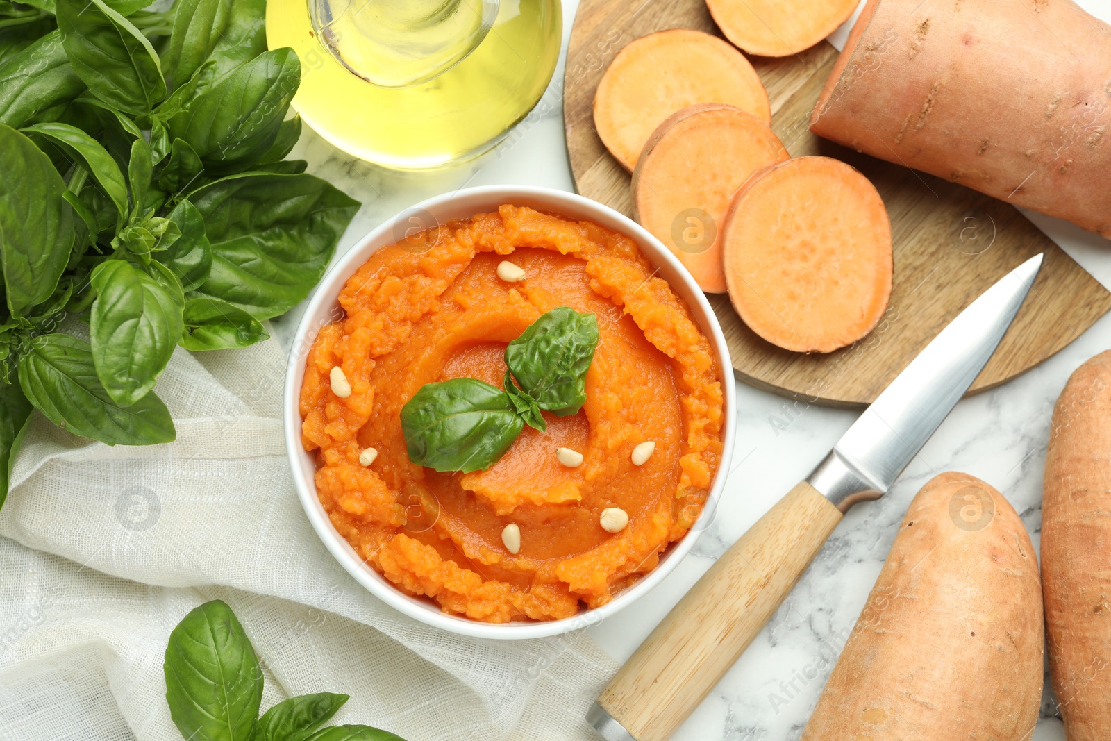 Photo of Tasty mashed sweet potato with basil, seeds in bowl and fresh vegetables on white marble table, flat lay