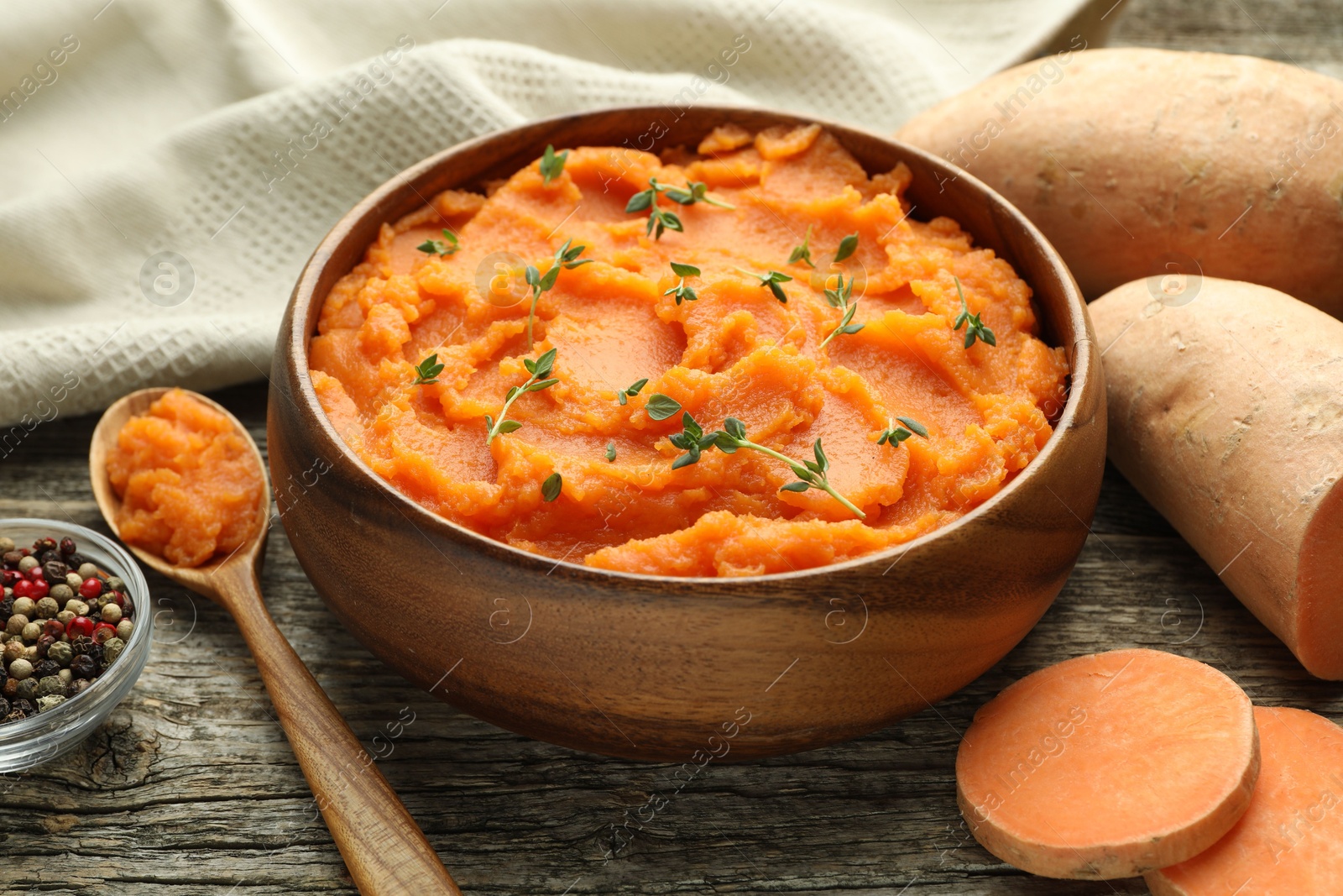 Photo of Tasty mashed sweet potato with thyme in bowl, fresh vegetables and spices on wooden table, closeup