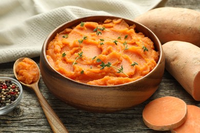 Photo of Tasty mashed sweet potato with thyme in bowl, fresh vegetables and spices on wooden table, closeup