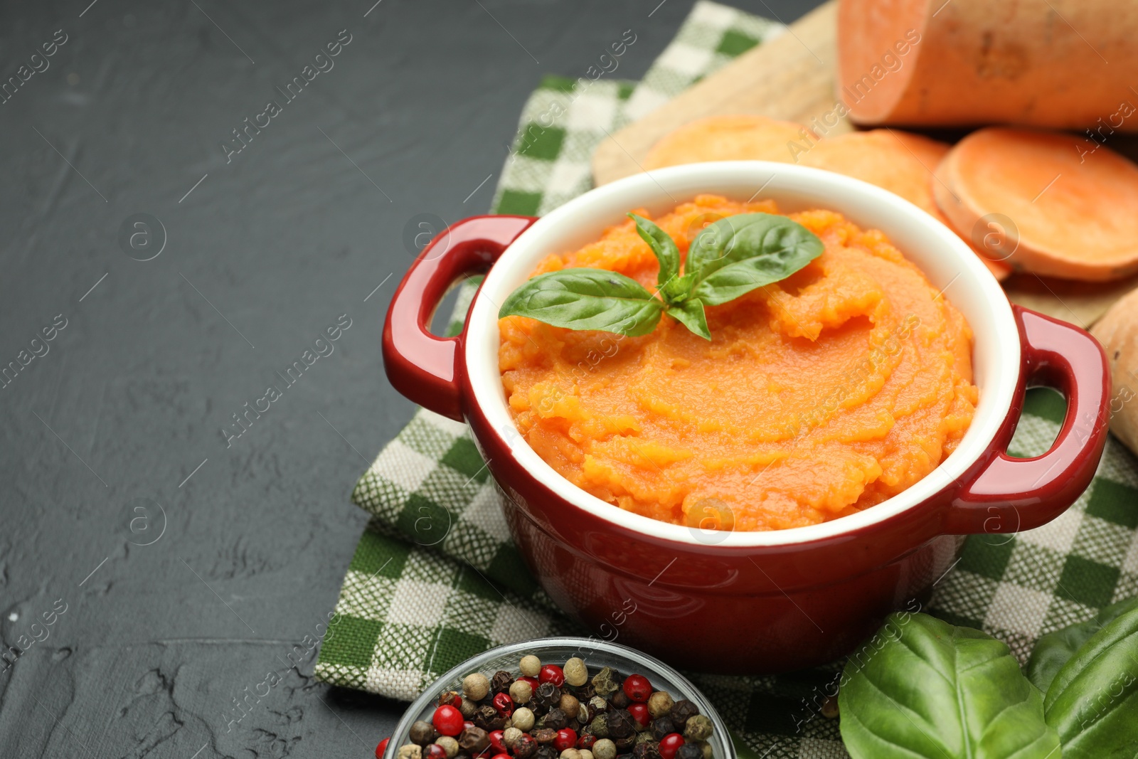 Photo of Tasty mashed sweet potato with basil in pot, cut vegetable and spices on dark textured table, closeup. Space for text