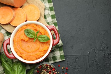 Photo of Tasty mashed sweet potato with basil in pot, cut vegetable and spices on dark textured table, flat lay. Space for text