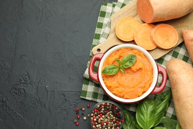 Photo of Tasty mashed sweet potato with basil in pot, cut vegetable and spices on dark textured table, flat lay. Space for text