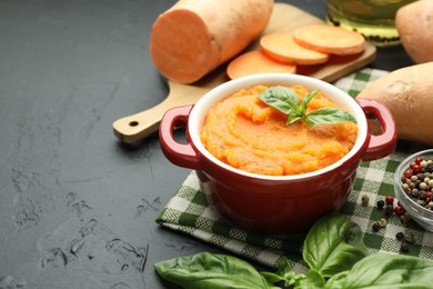 Photo of Tasty mashed sweet potato with basil in pot, fresh vegetables and spices on dark textured table, closeup