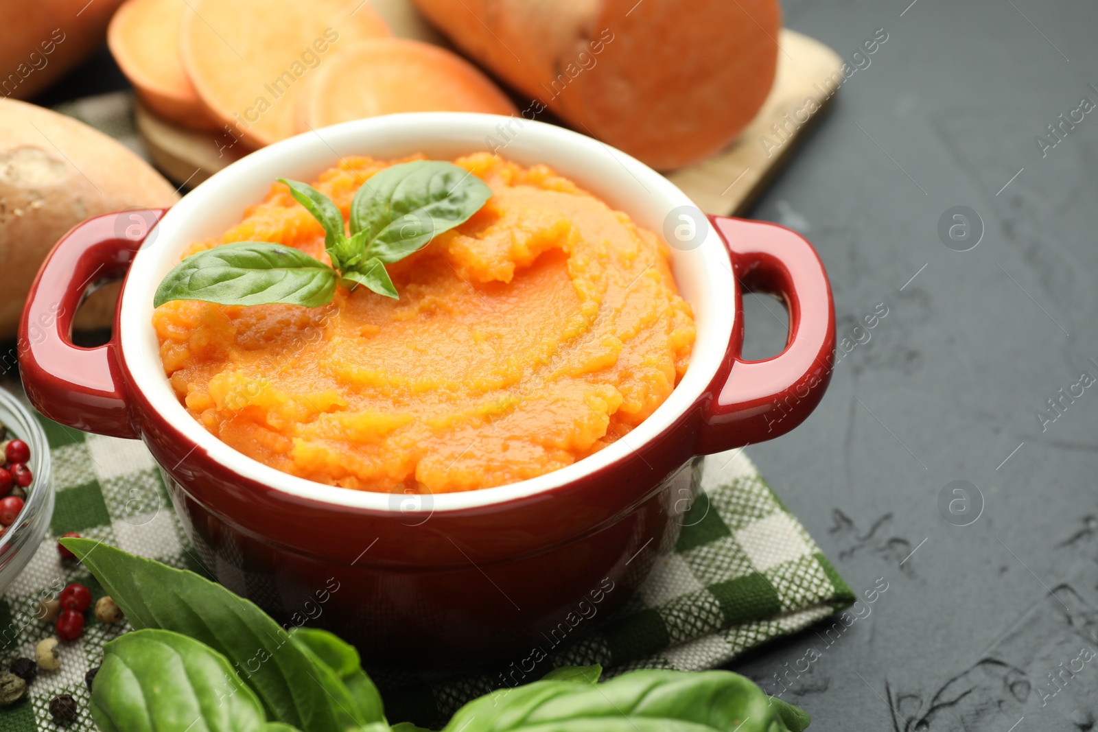 Photo of Tasty mashed sweet potato with basil in pot and fresh vegetables on dark textured table, closeup