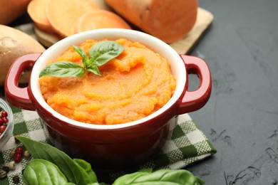 Photo of Tasty mashed sweet potato with basil in pot and fresh vegetables on dark textured table, closeup