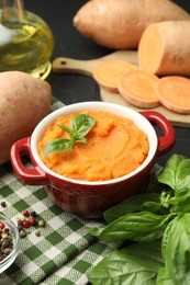 Photo of Tasty mashed sweet potato with basil in pot, fresh vegetables and spices on table, closeup