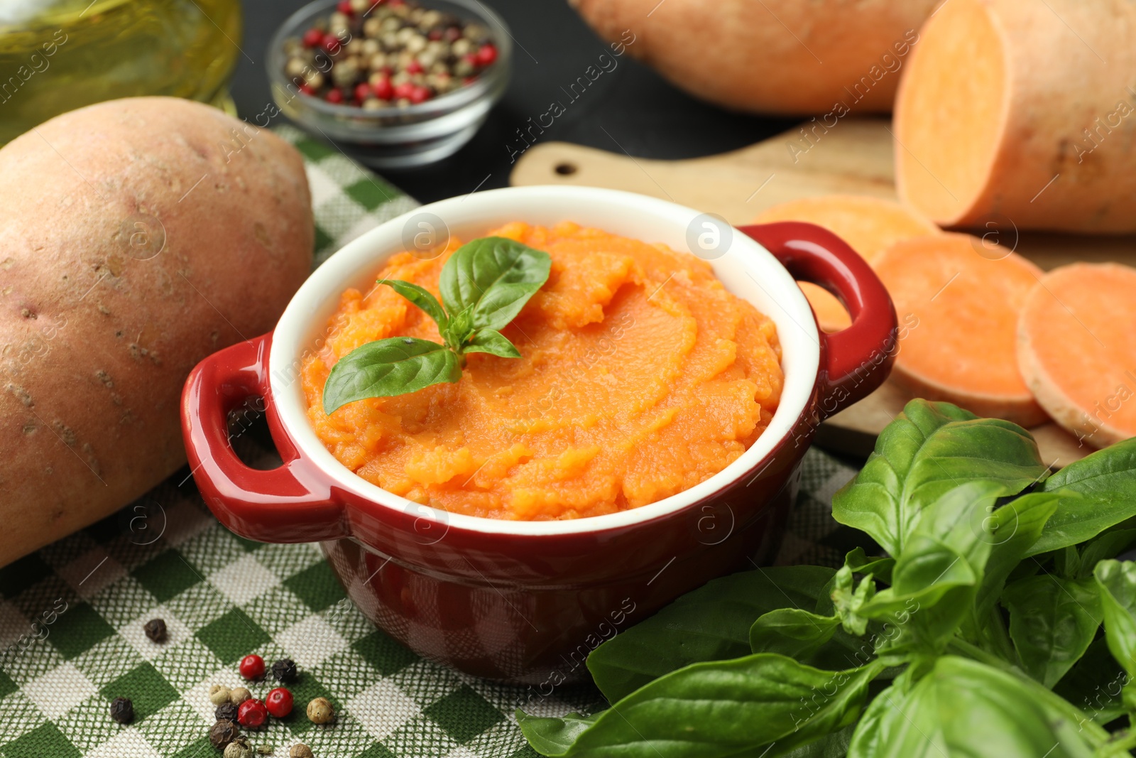 Photo of Tasty mashed sweet potato with basil in pot, fresh vegetables and spices on table, closeup