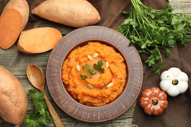 Photo of Tasty mashed sweet potato with pumpkin seeds in bowl, spoon, fresh vegetables and parsley on wooden table, flat lay