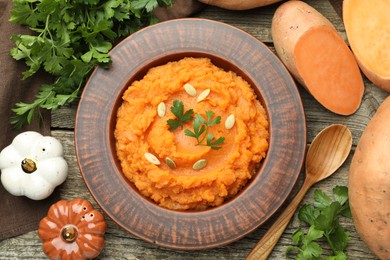 Photo of Tasty mashed sweet potato with pumpkin seeds in bowl, spoon, fresh vegetables and parsley on wooden table, flat lay