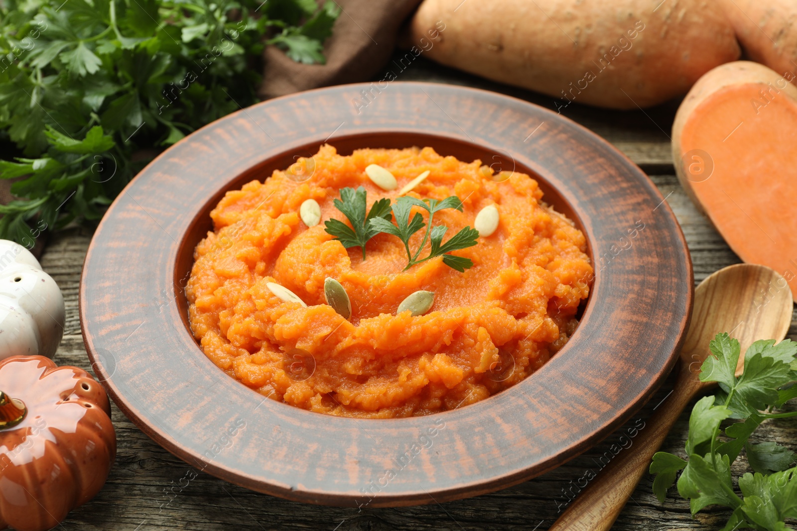 Photo of Tasty mashed sweet potato with pumpkin seeds in bowl, spoon, fresh vegetables and parsley on wooden table, closeup