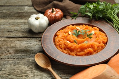 Photo of Tasty mashed sweet potato with pumpkin seeds in bowl, spoon, parsley and spices on wooden table, closeup