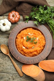 Photo of Tasty mashed sweet potato with pumpkin seeds in bowl, spoon, cut vegetable and parsley on wooden table, flat lay