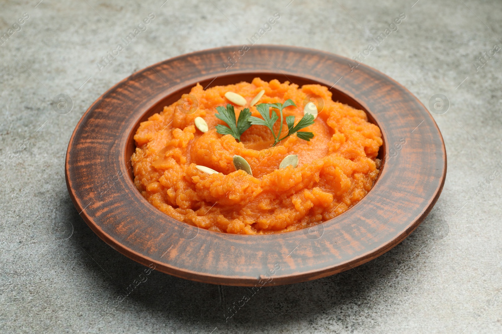 Photo of Tasty mashed sweet potato with pumpkin seeds and parsley in bowl on gray textured table, closeup