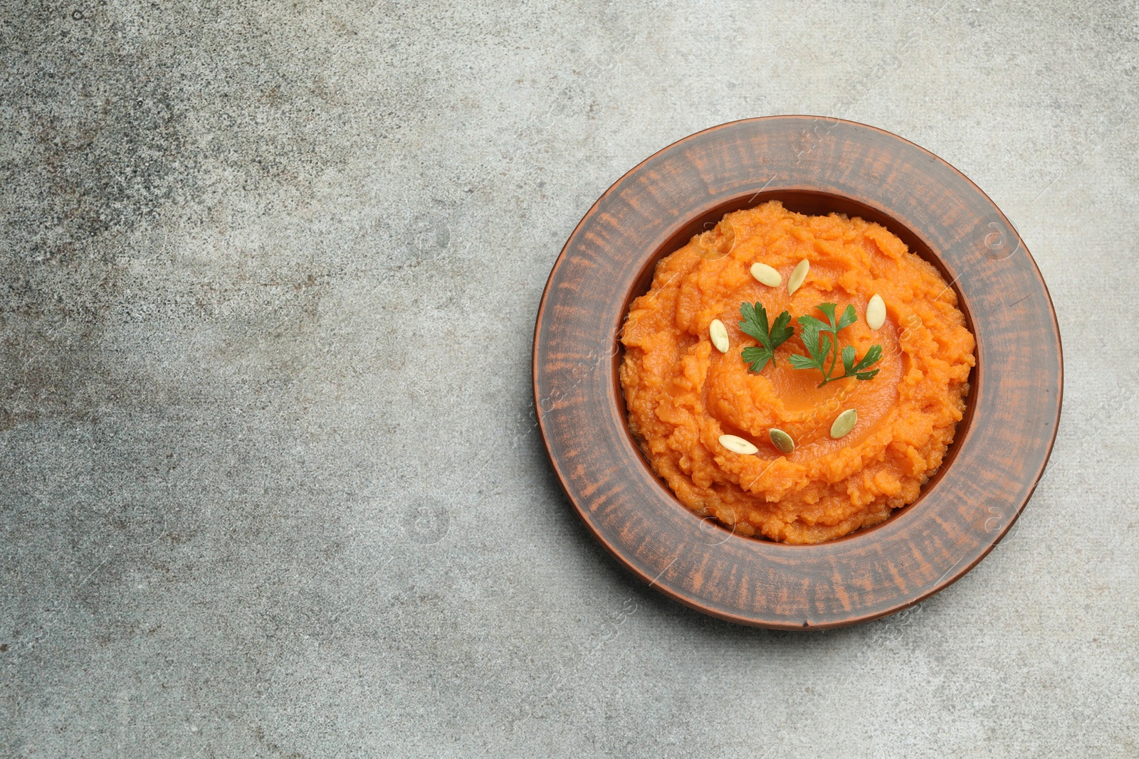 Photo of Tasty mashed sweet potato with pumpkin seeds and parsley in bowl on gray textured table, top view. Space for text