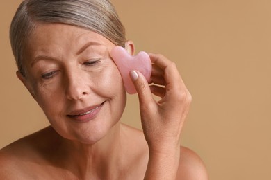 Beautiful woman doing facial massage with gua sha tool on beige background, closeup