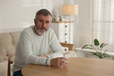 Photo of Senior man with glass of water at table indoors, space for text