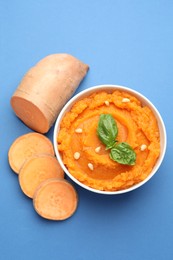 Photo of Tasty mashed sweet potato with basil, seeds in bowl and cut vegetable on blue background, flat lay