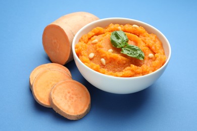 Photo of Tasty mashed sweet potato with basil, seeds in bowl and cut vegetable on blue background, closeup