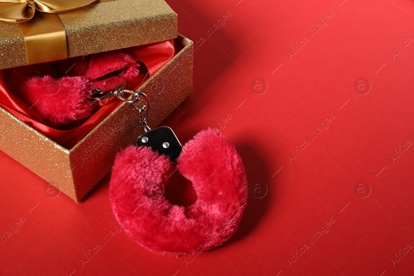 Photo of Bright fluffy handcuffs and gift box on red background, space for text