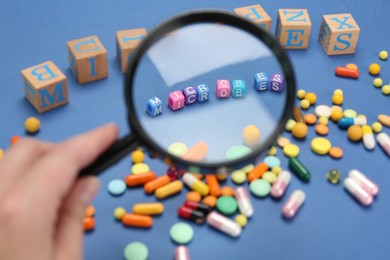 Photo of Woman holding magnifying glass over cubes with word Microbes on blue background, closeup