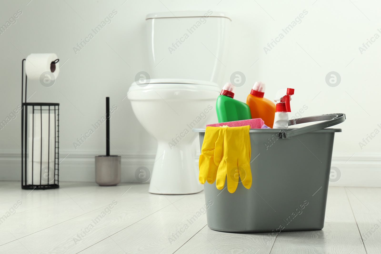 Photo of Bucket with different toilet cleaners and gloves on floor in bathroom