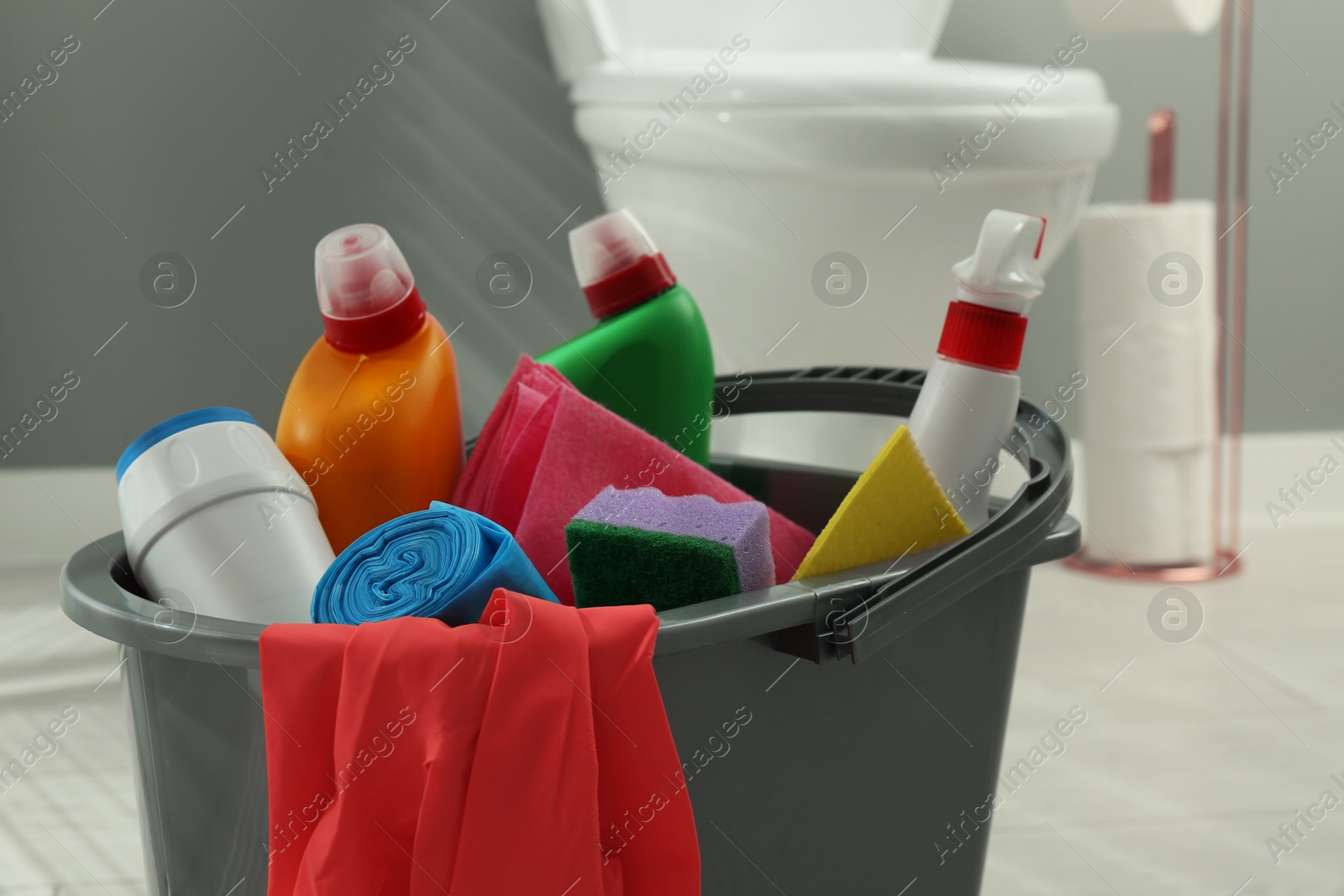 Photo of Bucket with different toilet cleaners, sponge, trash bags, rags and gloves in bathroom, closeup