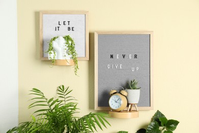 Photo of Letter boards with phrases Let It Be, Never Give Up, alarm clock and plants on beige wall