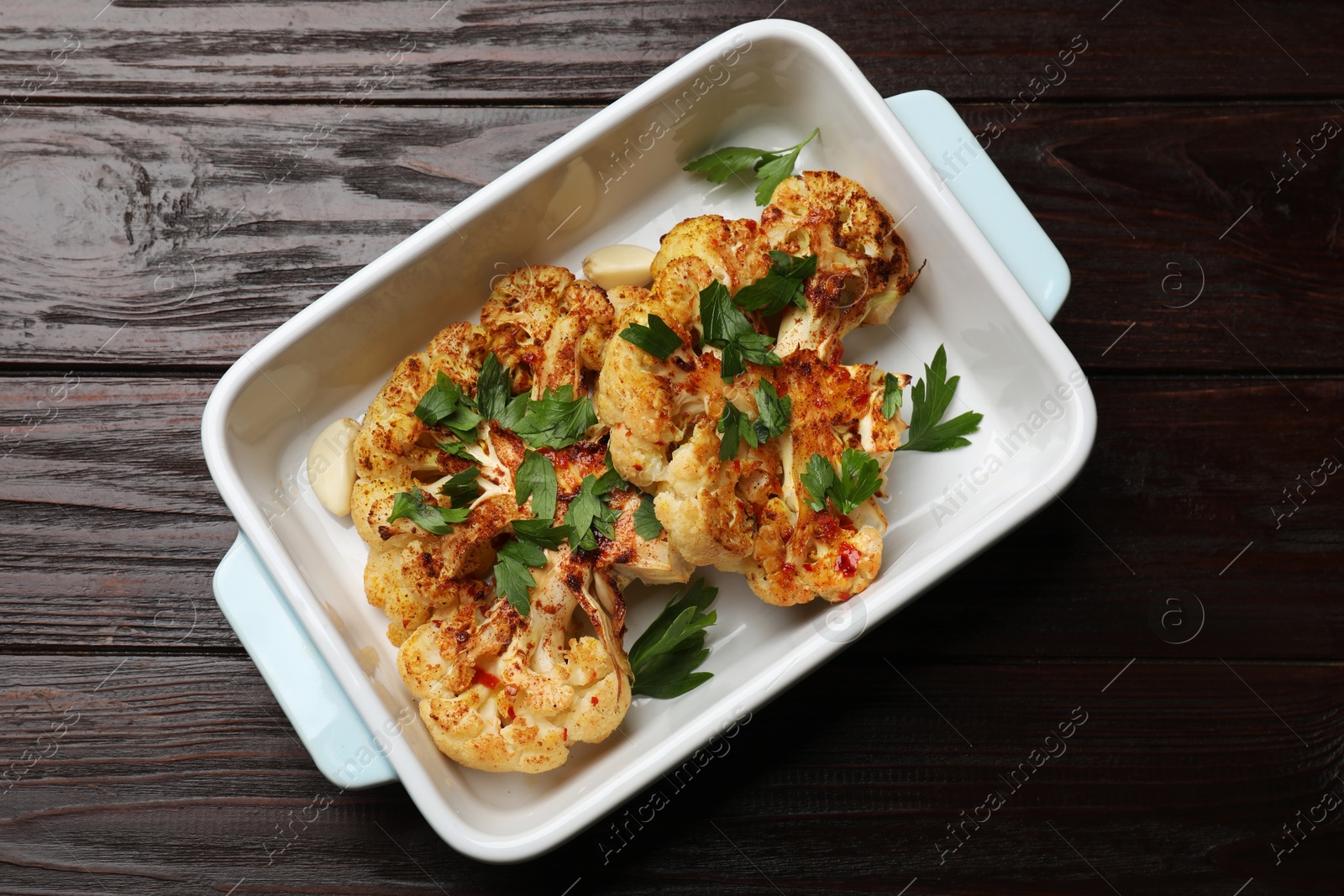 Photo of Tasty cauliflower steaks and parsley in baking dish on wooden table, top view